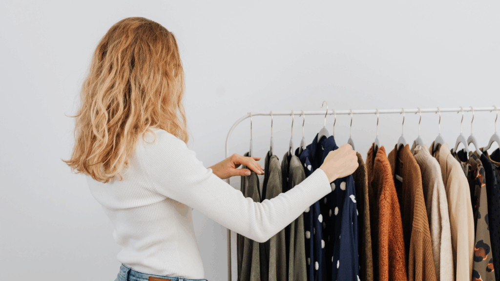 A woman with blonde hair looks through clothes on a rack against a plain white wall.