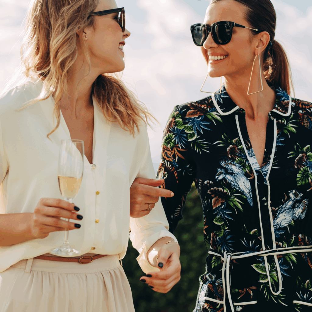 Two women in sunglasses walk outdoors, smiling and holding champagne glasses, dressed in stylish casual outfits.
