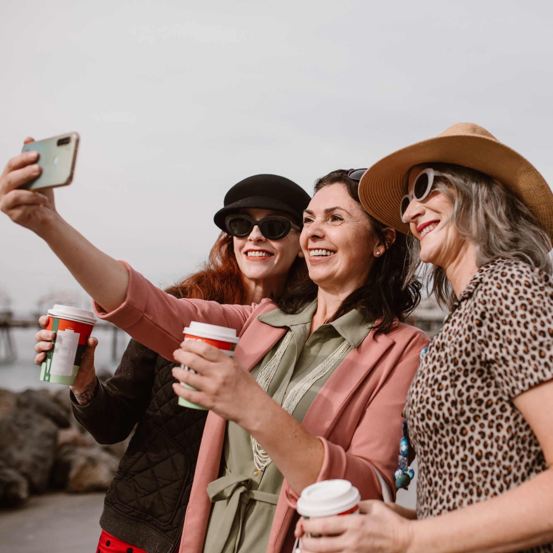 Three women smiling and taking a selfie outdoors while holding coffee cups.