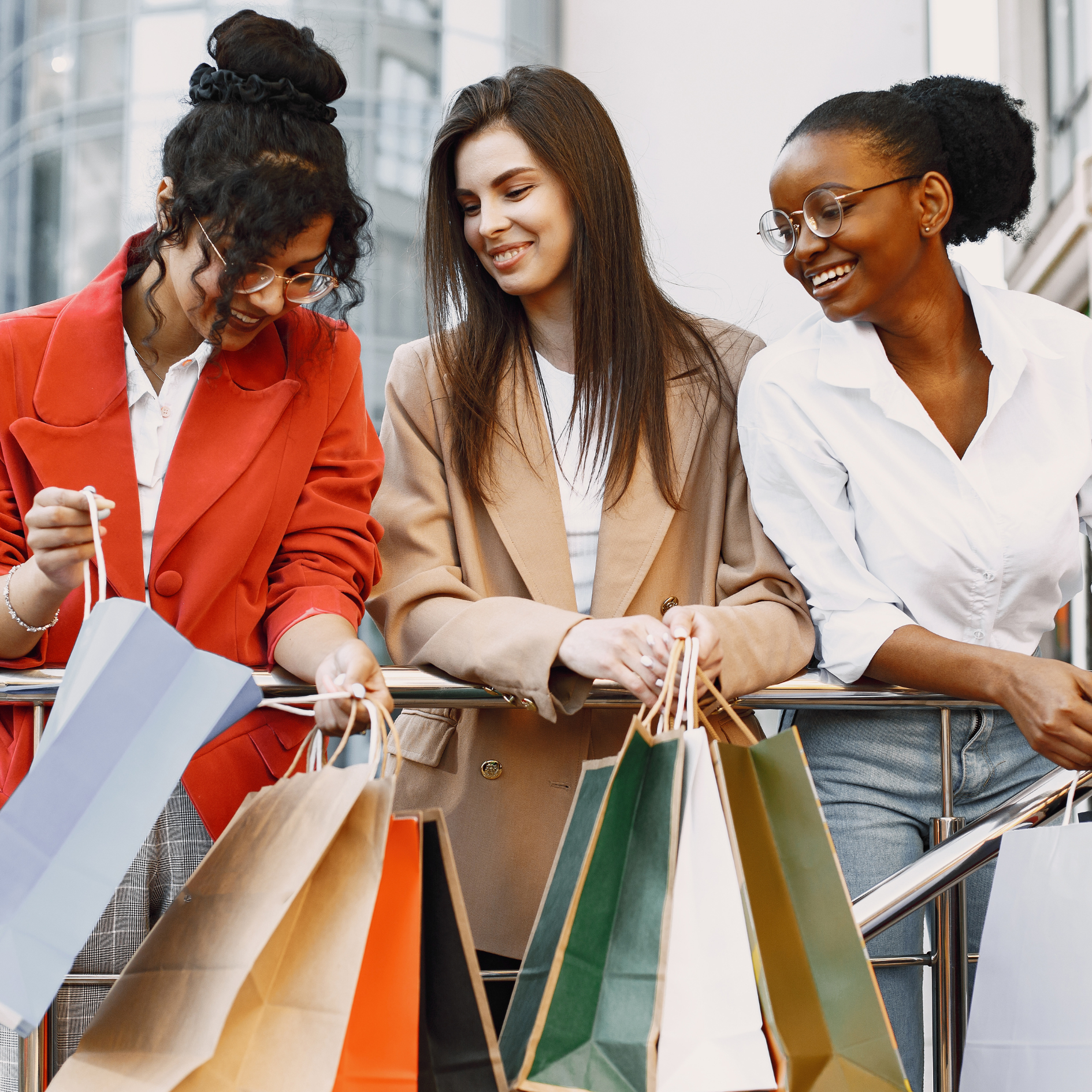 Three women stand outdoors, smiling and holding multiple shopping bags over a railing.