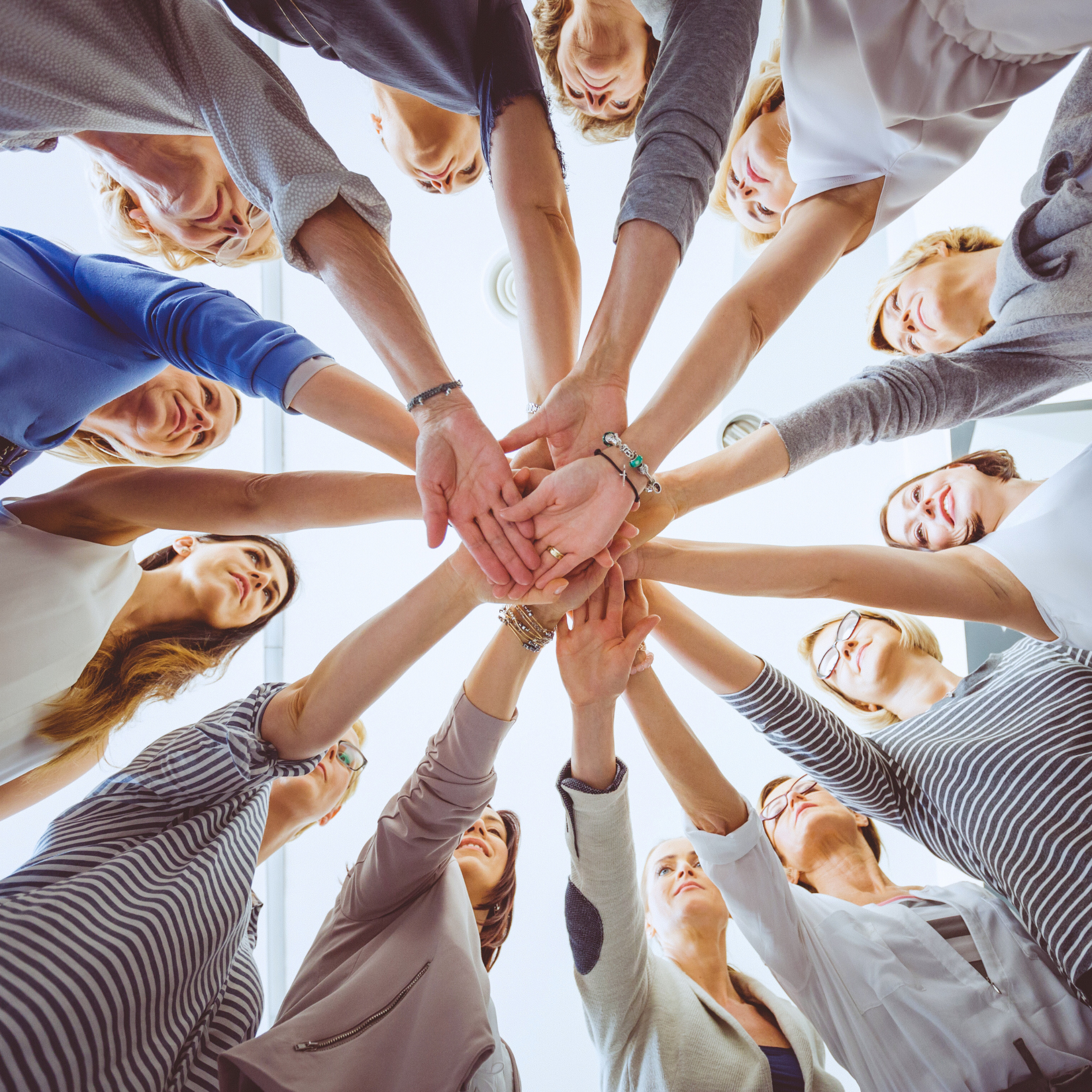 Group of people standing in a circle, reaching into the center to stack their hands together in a gesture of unity.