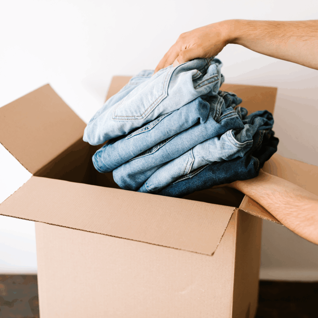 A person places a stack of folded denim jeans into an open cardboard box.
